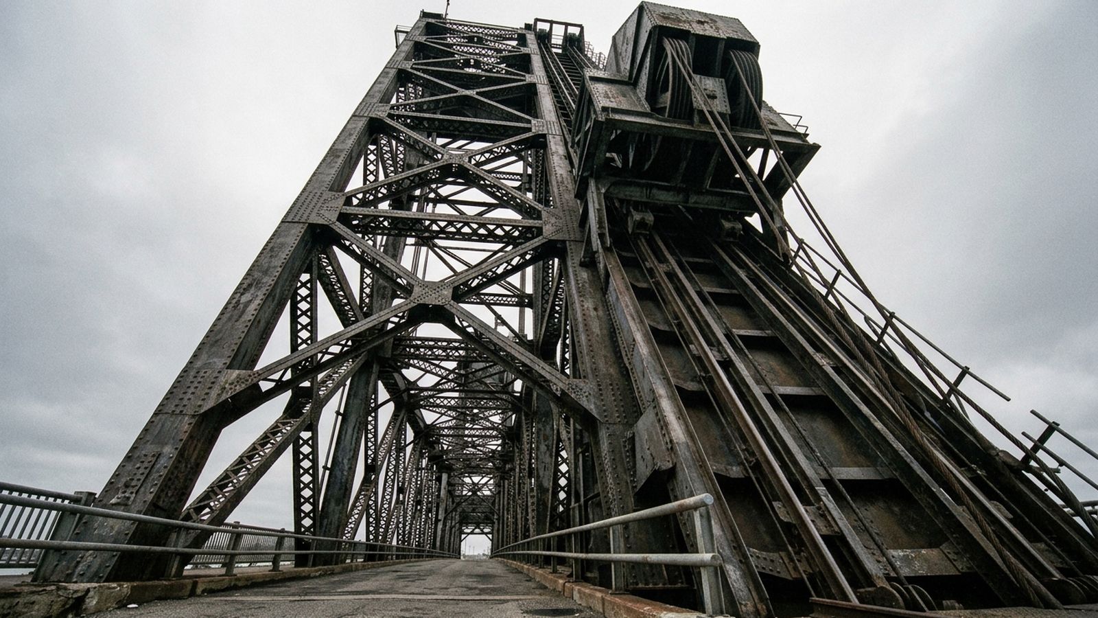 Looking up the steel lattice leg of a vertical lift bridge tower from the sidewalk beneath it, the sheave housing visible at the top and the counterweight guide track running down the inside of the tower, flat gray daylight, no signage or lettering