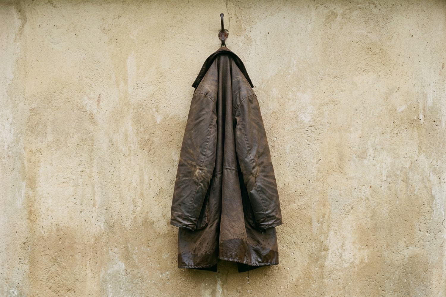 A worn oilskin coat on an iron hook against a plain wall, still wet at the hem.