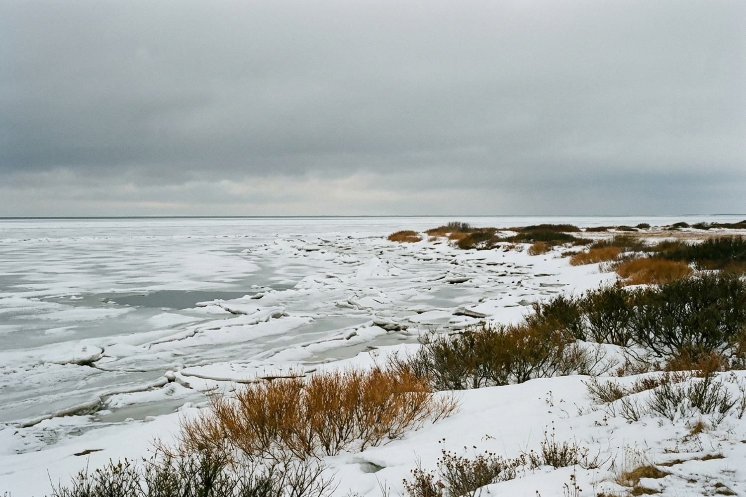 A flat frozen shoreline of tidal ice and drifted snow meeting a gray sky.