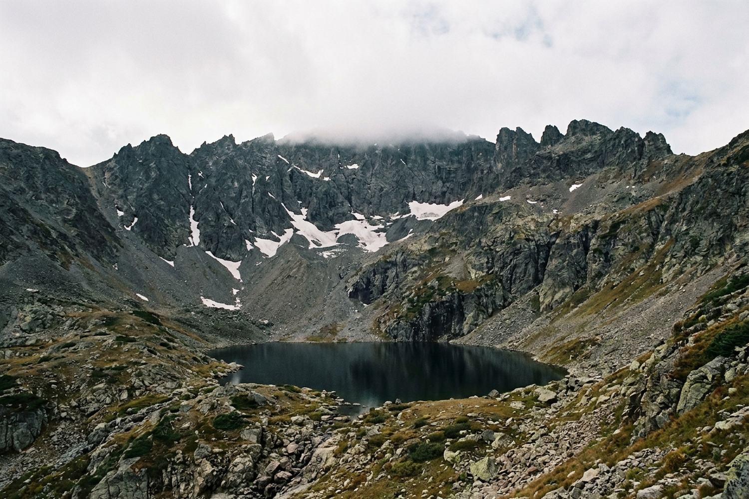A rocky ridge above a small dark lake, old snow in the gullies, cloud below the tops.