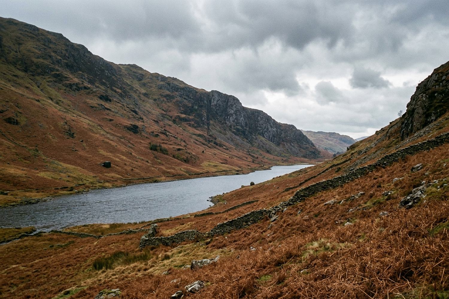 A bracken covered mountainside above a long narrow lake under torn gray cloud.